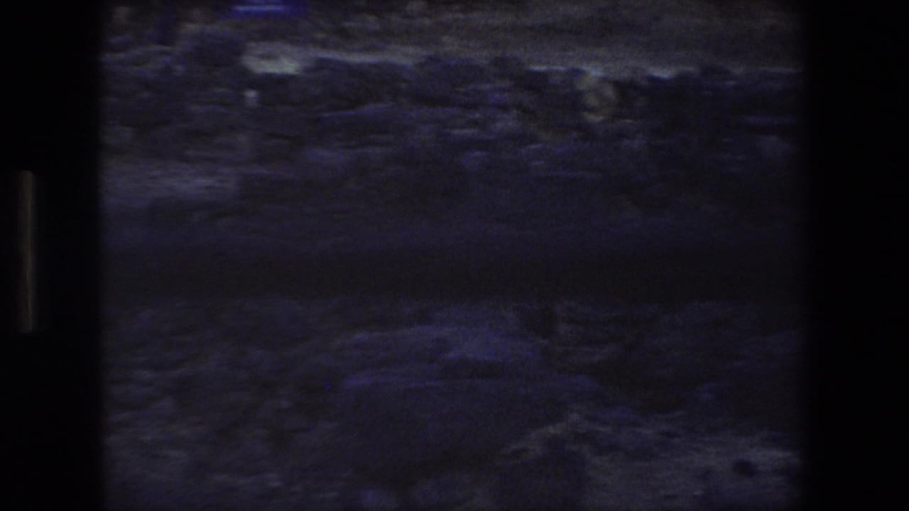 Final 8mm frame showing a dim handheld view of rocky ground and low ruin stones at the Tel Aviv site, framed by dark film edges.