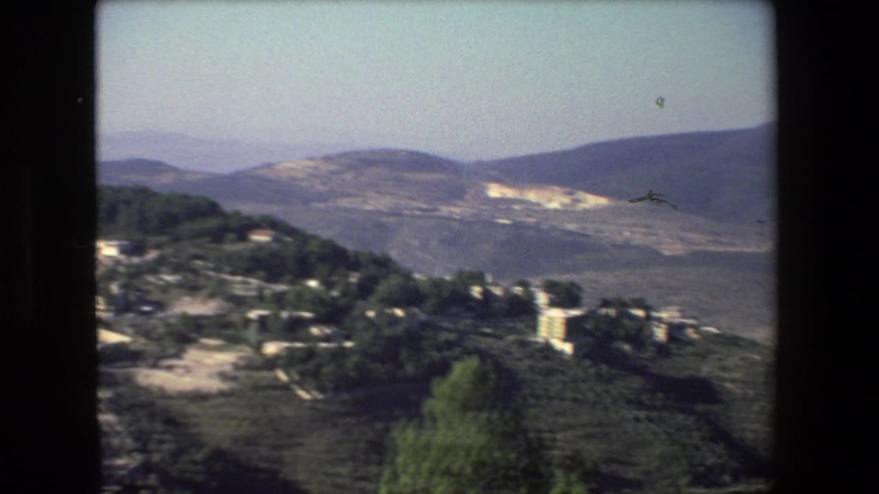 Soft 8mm frame from the 1972 Israel-labeled footage showing hillside buildings, dark green trees, a pale road, and layered mountains under a washed sky.