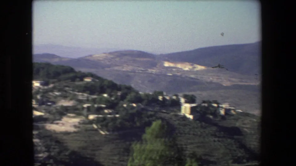Soft 8mm frame from the 1972 Israel-labeled footage showing hillside buildings, dark green trees, a pale road, and layered mountains under a washed sky.