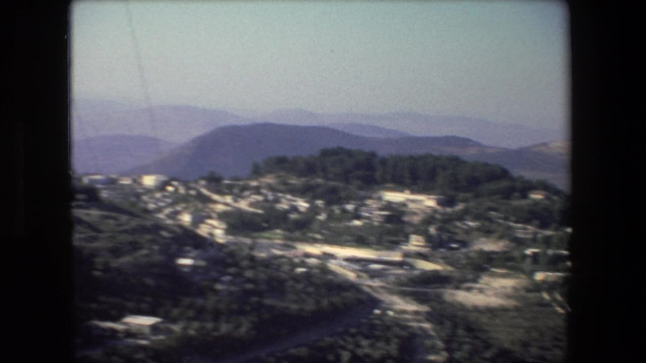Elevated 8mm view from the Israel-labeled 1972 clip showing a cluster of buildings, wooded slopes, a road, and layered blue-purple hills in the distance.