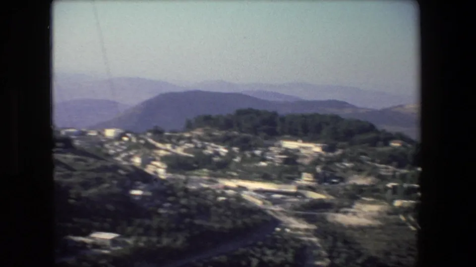 Elevated 8mm view from the Israel-labeled 1972 clip showing a cluster of buildings, wooded slopes, a road, and layered blue-purple hills in the distance.