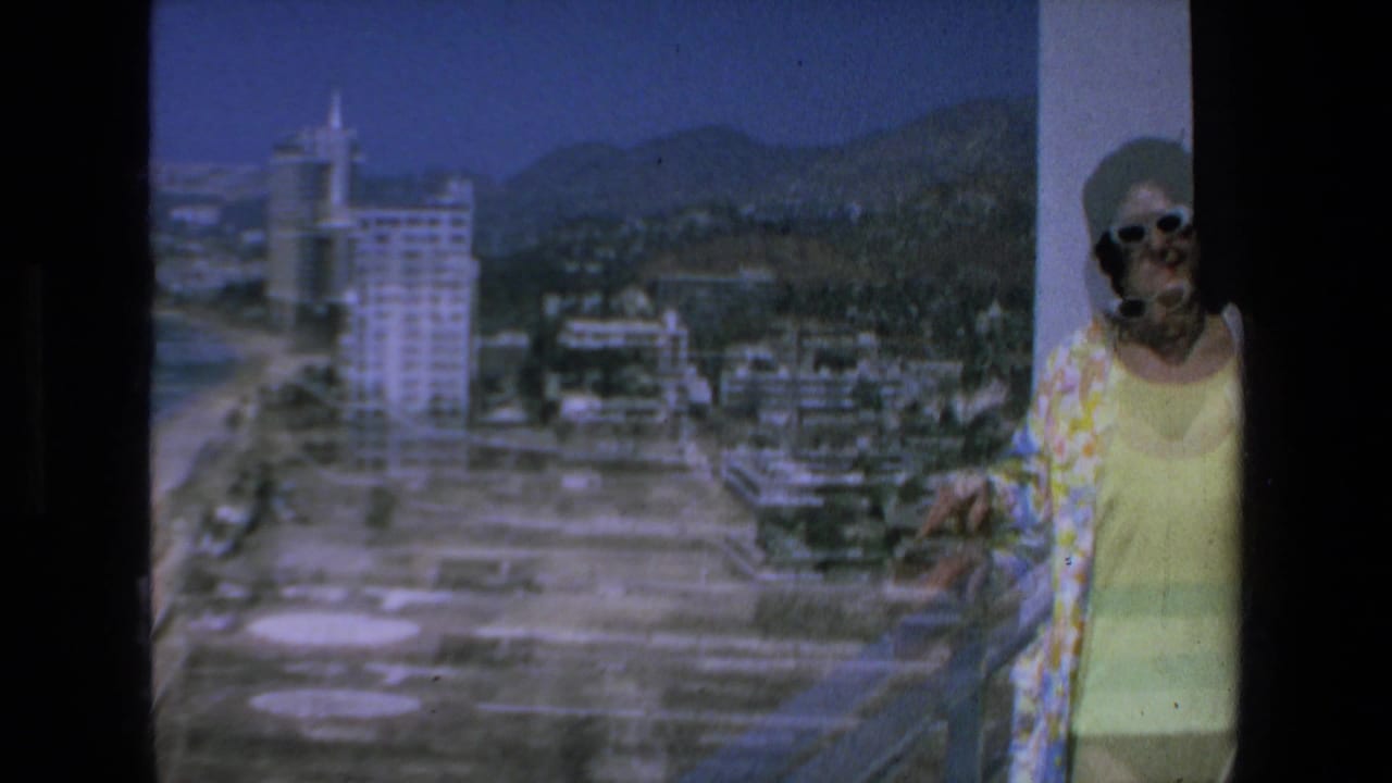 In 1972 Mazatlan, Mexico, a woman in white sunglasses and a yellow outfit stands near a balcony railing with beachfront hotels, shoreline, and mountains behind her.