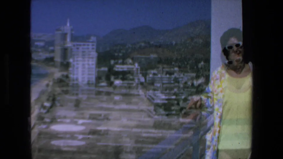 In 1972 Mazatlan, Mexico, a woman in white sunglasses and a yellow outfit stands near a balcony railing with beachfront hotels, shoreline, and mountains behind her.
