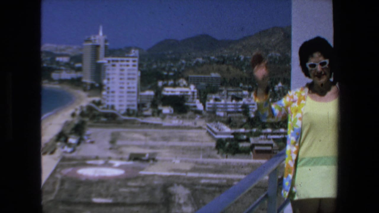 Woman waves from a high balcony with beach, hotels, and mountains in the background.