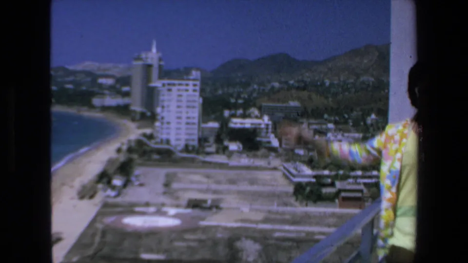 The woman is partly cropped at the edge of the 1972 Mazatlan balcony view while the camera emphasizes the beach curve, high-rise buildings, city area, and distant hills.