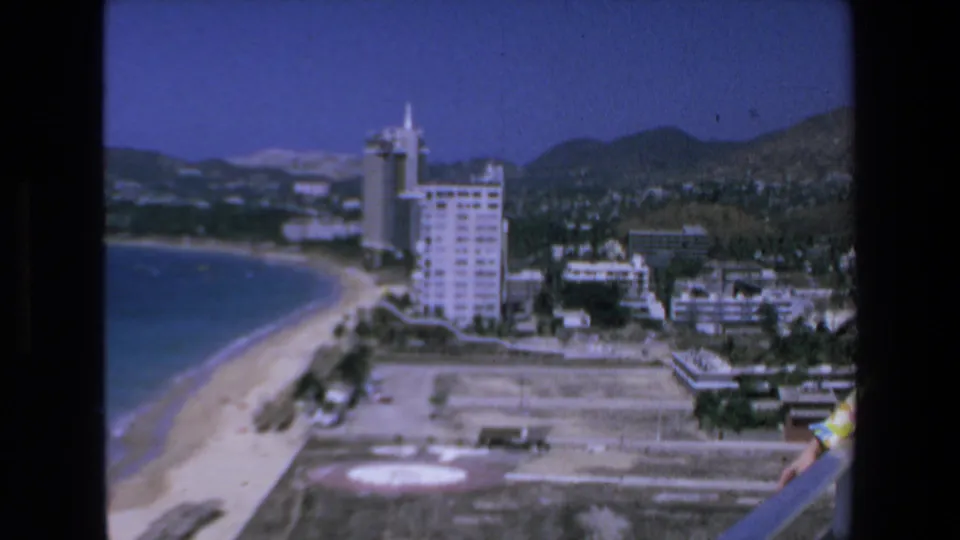 A wide 8mm view from a balcony shows the Mazatlan coastline, beach, hotel buildings, city development, and mountains, with the woman only partly visible at the right edge.