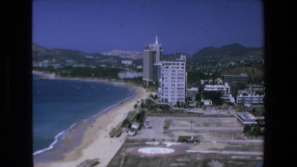 1972 Mazatlan home-movie frame showing calm blue water, a sandy shoreline, tall coastal hotels, a beachfront helipad, and hills under clear sky.