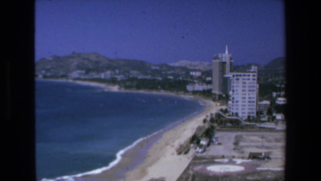 Ocean curves toward beachfront buildings with hills behind the shoreline.