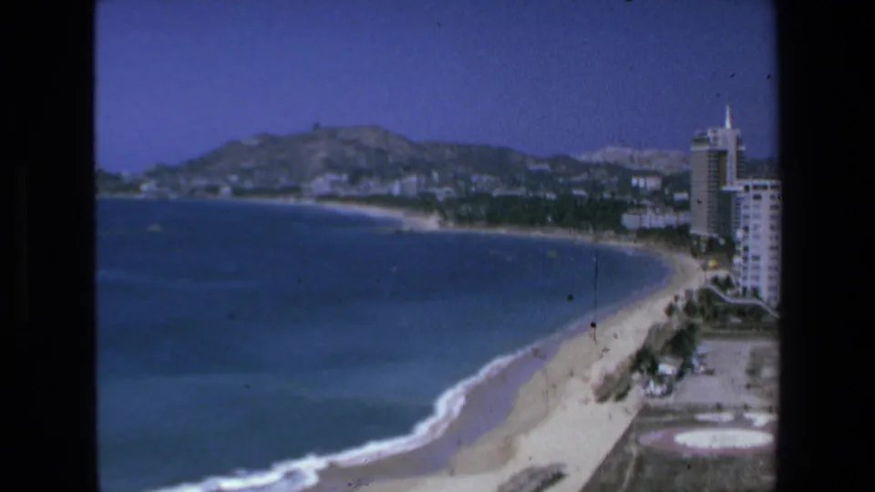 1972 Mazatlan frame looking along the beach and ocean toward hills, with tall shoreline buildings partly visible at the right edge.