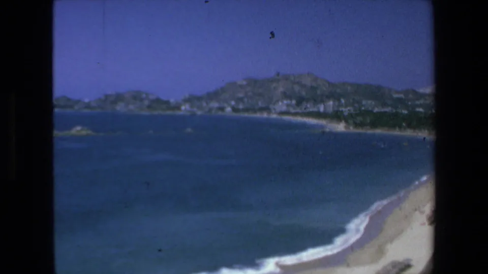 1972 Mazatlan home-movie frame showing a wider ocean view, curved beach, distant coastal buildings, and hills under a clear blue sky.