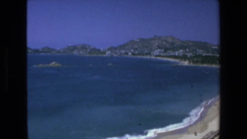 1972 8mm home-movie view of Mazatlan coastline with blue ocean water, a curved sandy beach, distant hillside buildings, and visible film-frame edges.