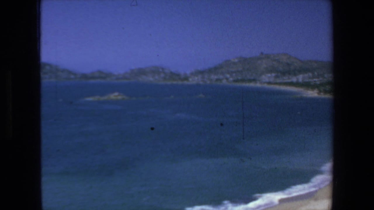 Wide ocean view with distant hills, rocky islets, and a strip of beach at the right edge.