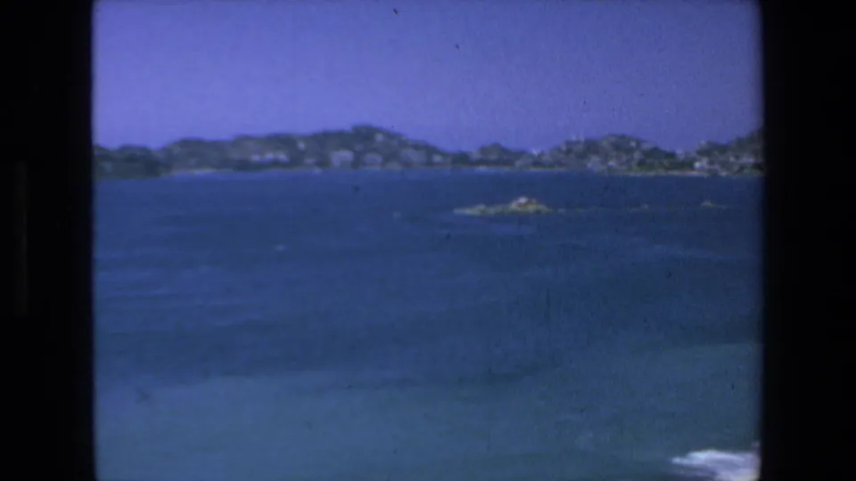 Soft 8mm frame from Mazatlan showing open blue water, small rocky islets, distant hills, and faint shoreline buildings under a clear sky.