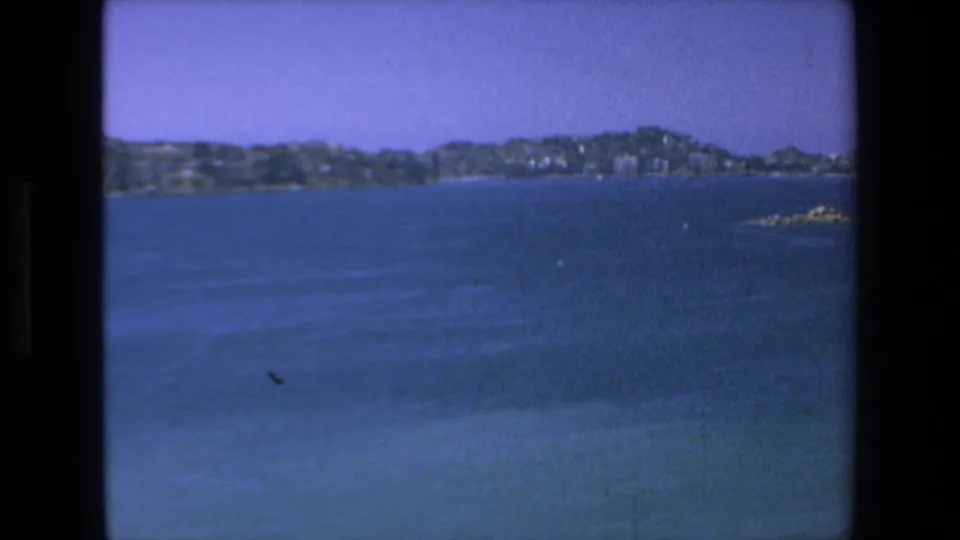 1972 8mm view across Mazatlan Bay toward low hills and shoreline buildings, with blue water filling most of the frame and light film wear visible.