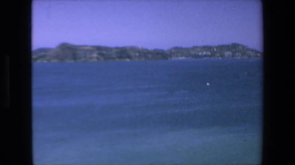 1972 Mazatlan home-movie frame showing calm blue water and a distant tree-covered island or shoreline in soft 8mm color.