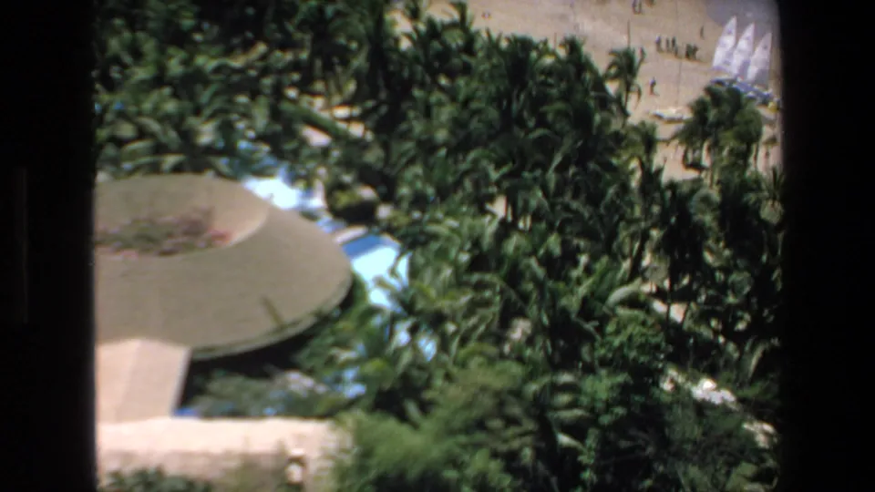 1972 Mazatlan home-movie frame looking over palms and a circular seaside structure toward a sandy beach with white sailboats and distant people.