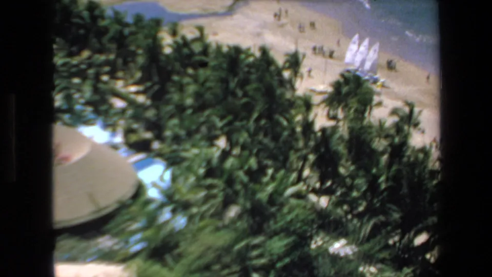 Soft 8mm 1972 Mazatlan frame of a palm-lined beach area with small beachgoers, white sailboats on the sand, and water at the edge of the frame.