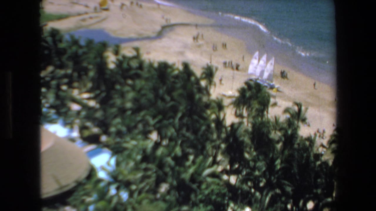 1972 Mazatlan 8mm frame showing a resort beach from above, with palms in the foreground, white sailboats on the sand, scattered people, and ocean surf beyond.