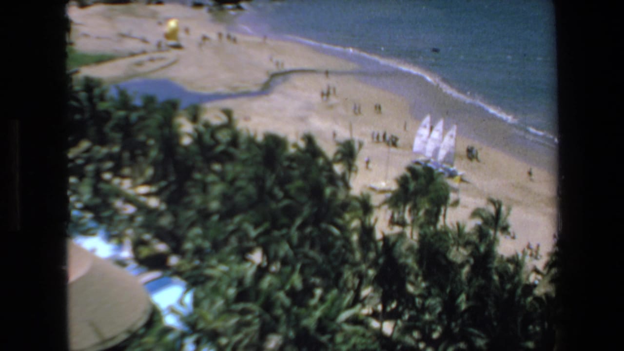 Elevated 1972 Mazatlan beach view with dense palms in the foreground, sailboats near the shoreline, small figures on the sand, and blue water at the top of frame.