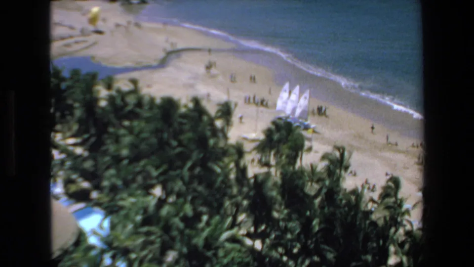 Soft 8mm frame from Mazatlan in 1972 showing palms, beachgoers, white sailboats, and a narrow line of surf, filmed from an elevated resort-side position.