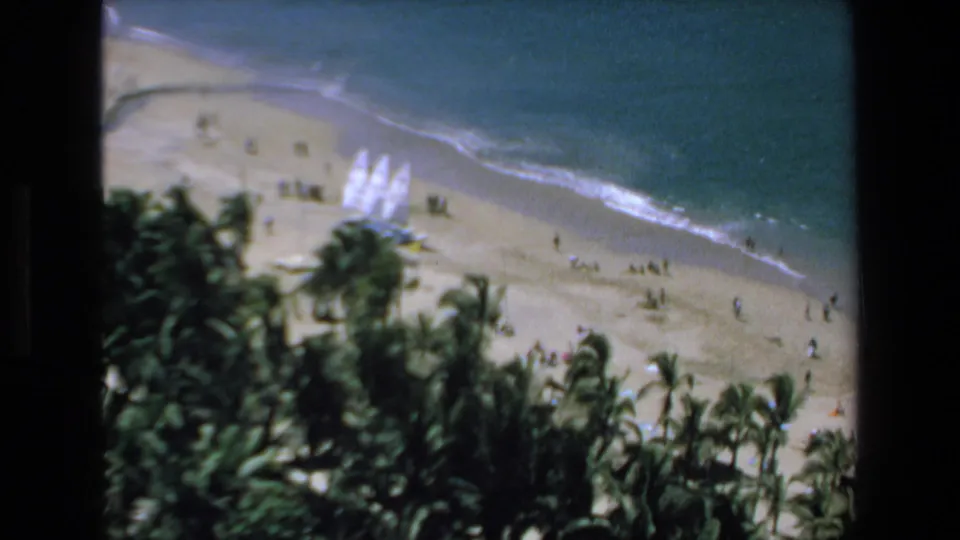 Later frame of the 1972 Mazatlan beach scene with palms below, people spread across the sand, several white sailboats grouped near shore, and gentle surf in the background.