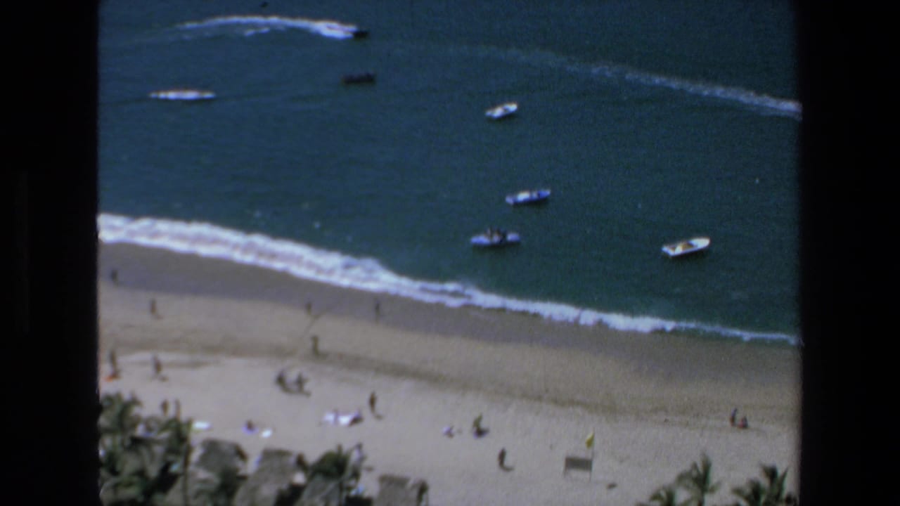 1972 Mazatlan home-movie frame looking down at a beach with scattered visitors, surf, palm tops, and small boats floating offshore.
