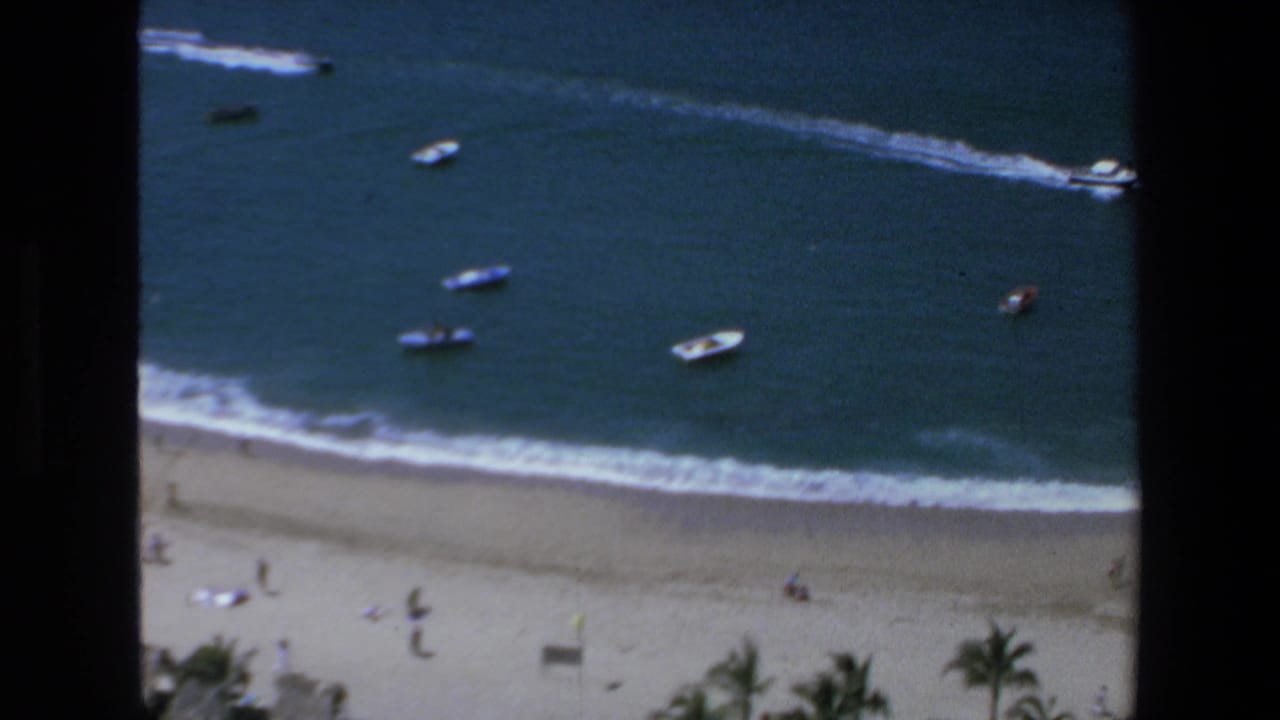 Elevated 1972 Mazatlan coastal view with small boats beyond the surf and distant beachgoers on the sand below.