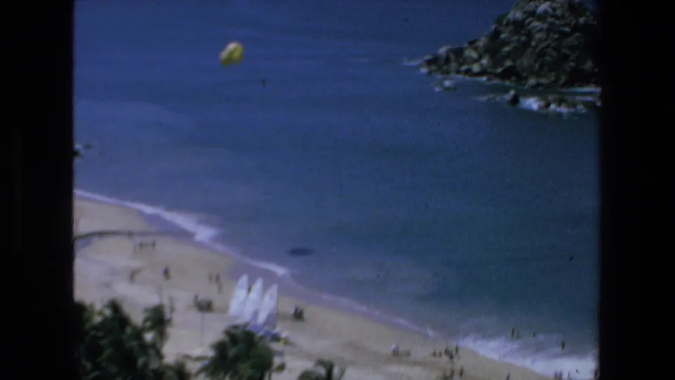 1972 Mazatlan 8mm frame showing a yellow parasail above blue water near a beach, rocky shoreline, palm trees, and small distant figures.