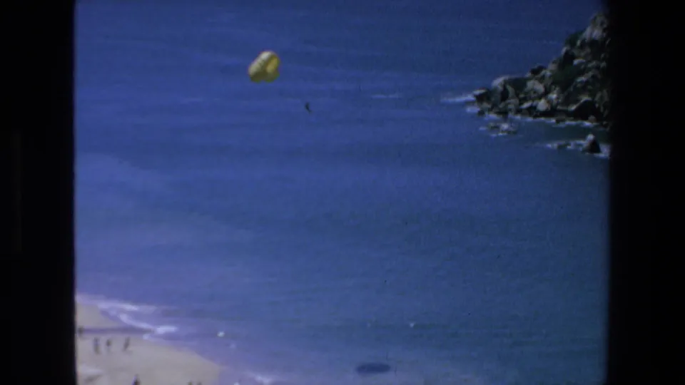 Final 1972 Mazatlan frame with a yellow parasail suspended over open water, a rocky coast at right, and a beach edge with distant people below.