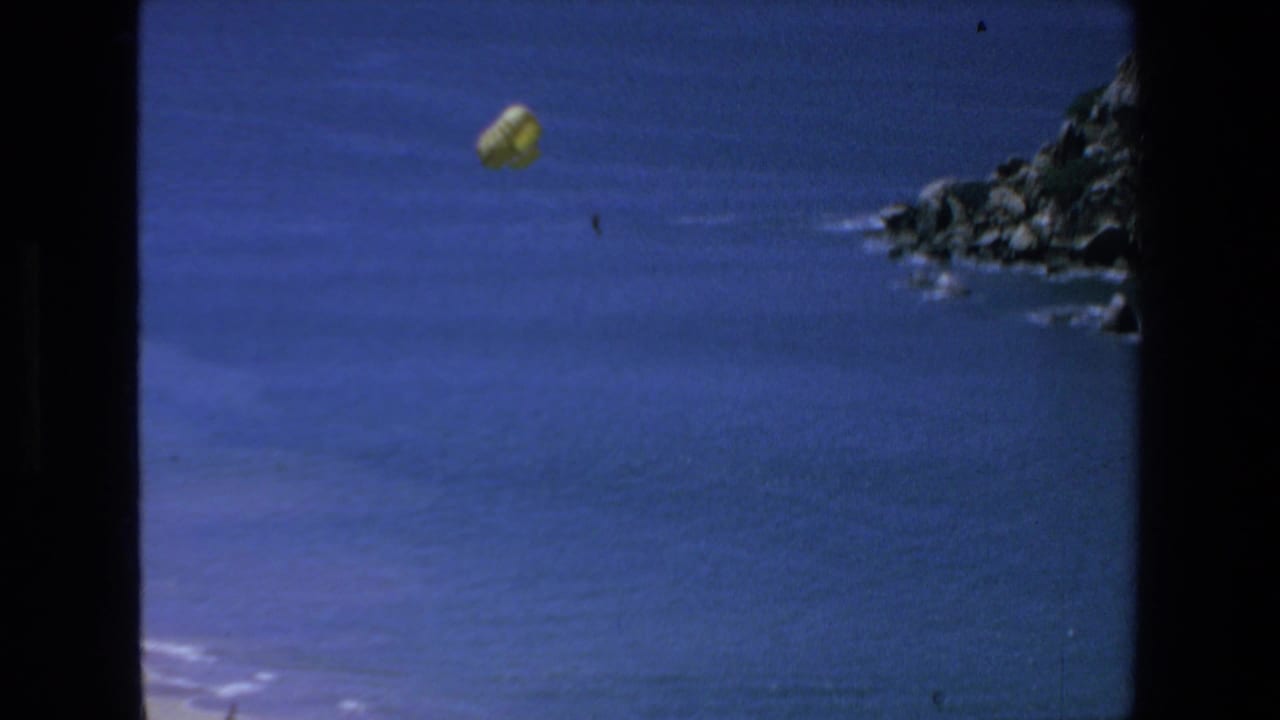 A yellow parachute floats over the blue ocean near a rocky Mazatlan coastline in 1972 8mm home-movie footage.
