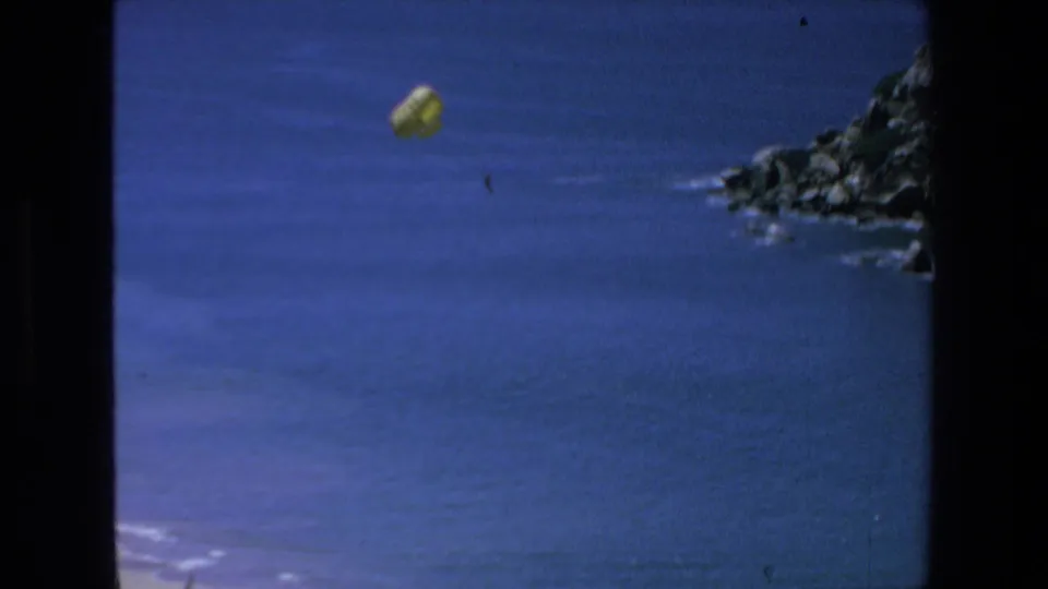 A yellow parachute floats over the blue ocean near a rocky Mazatlan coastline in 1972 8mm home-movie footage.