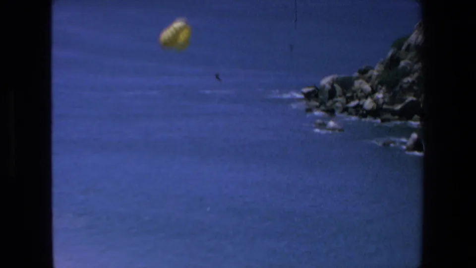 A distant yellow parachute appears over open Pacific water, with dark rocky coastline visible at the right edge of the 1972 Mazatlan frame.