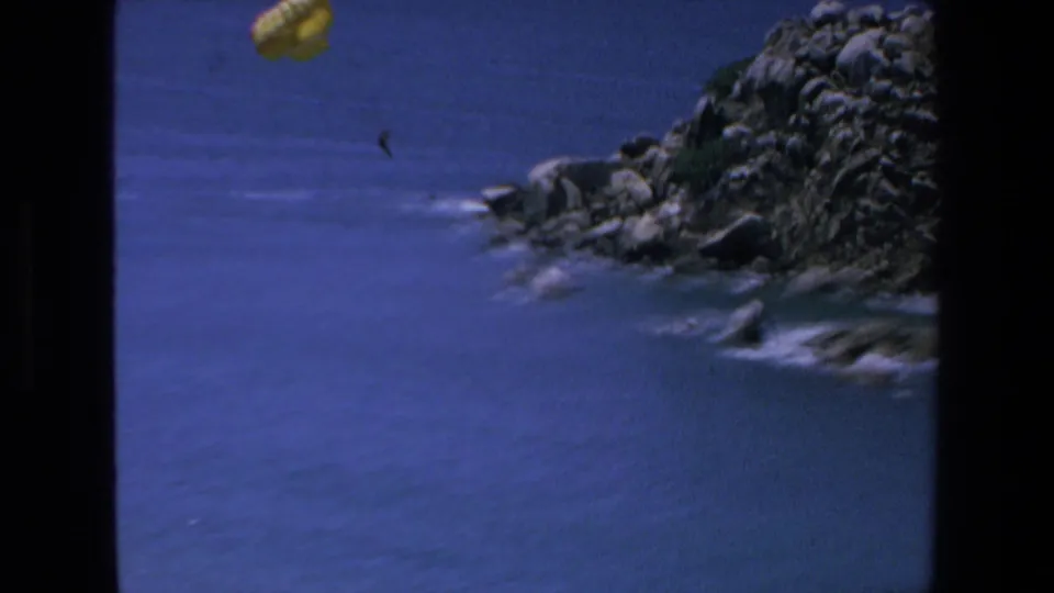 The yellow parachute moves near the upper frame above blue coastal water, with rocky shoreline and small surf visible below in Mazatlan home-movie footage.