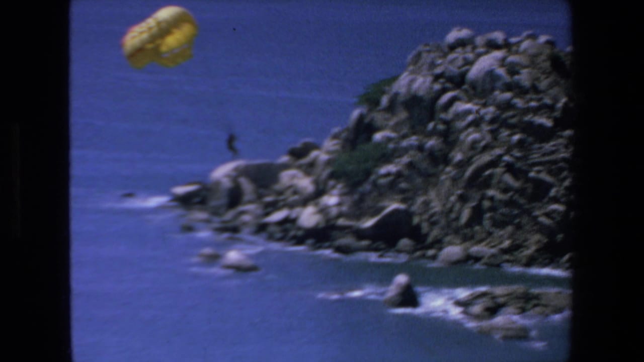 In Mazatlan, Mexico, 1972, a yellow parachute carries a distant person over blue water beside rocks and surf in soft 8mm footage.