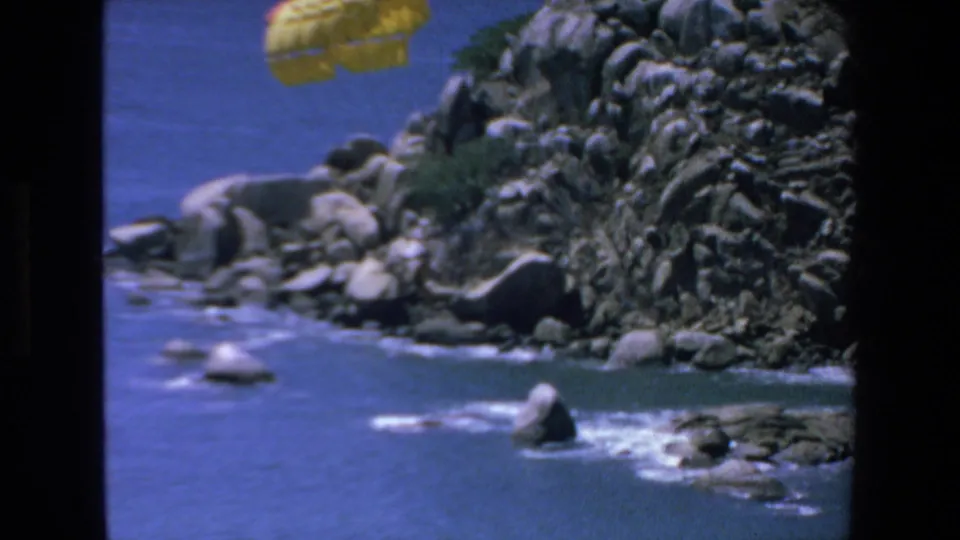 The yellow parachute drifts across the upper frame above the rugged Mazatlan shoreline, with blue water and white surf below.
