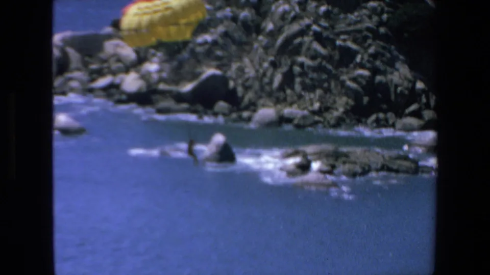A yellow parachute with a distant person beneath it descends over blue water near the rocky Mazatlan coast in a brief 1972 8mm travel scene.