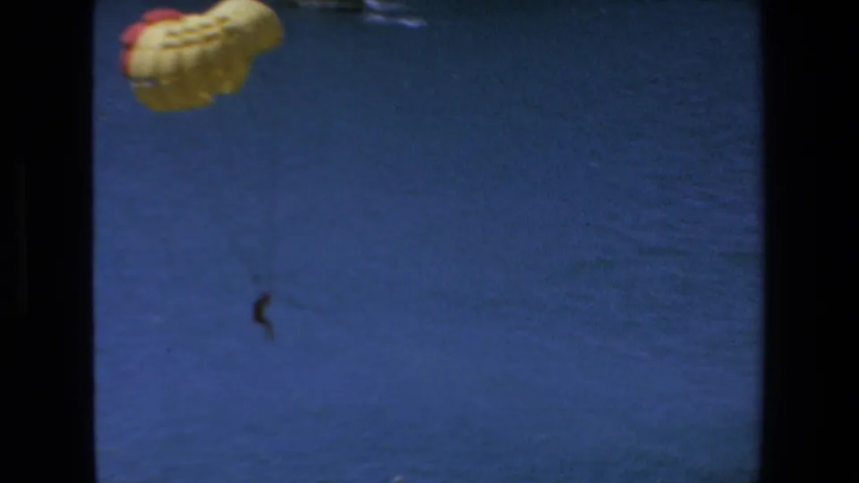 1972 Mazatlan 8mm frame showing a cropped yellow parasail at upper left and a small parasailer hanging below over rippled ocean water.