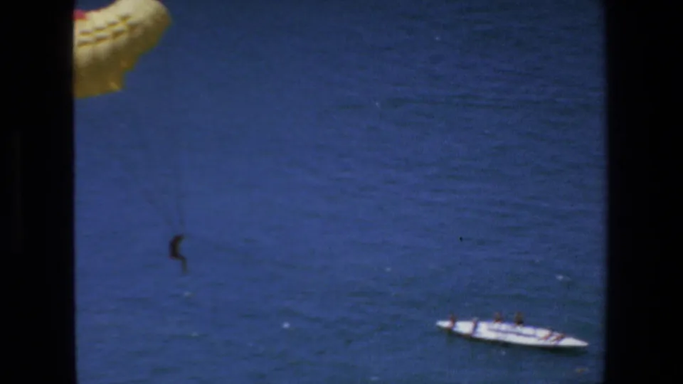 1972 Mazatlan home-movie frame showing the parasailer still suspended over the ocean, with part of the yellow canopy at top left and a small boat carrying people near the lower right.