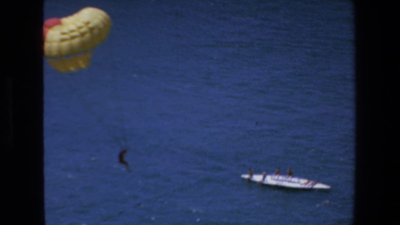 1972 Mazatlan home-movie frame showing a yellow and red parasail above the sea with a distant person suspended and a white tow boat below.
