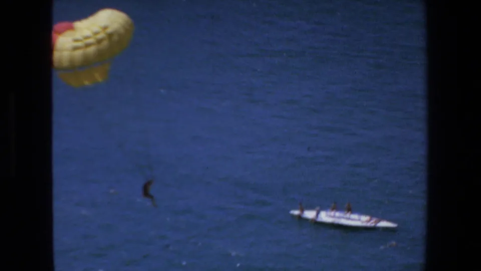 1972 Mazatlan home-movie frame showing a yellow and red parasail above the sea with a distant person suspended and a white tow boat below.