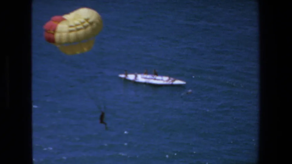 1972 Mazatlan home-movie frame showing the parasail closer to the left side of the frame, with the white tow boat and a dark suspended figure over the water.