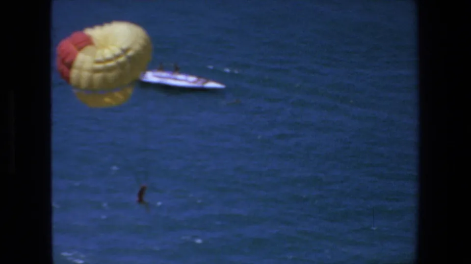 1972 Mazatlan frame showing the yellow and red parasail near the tow boat, with the suspended person lower over the open sea.