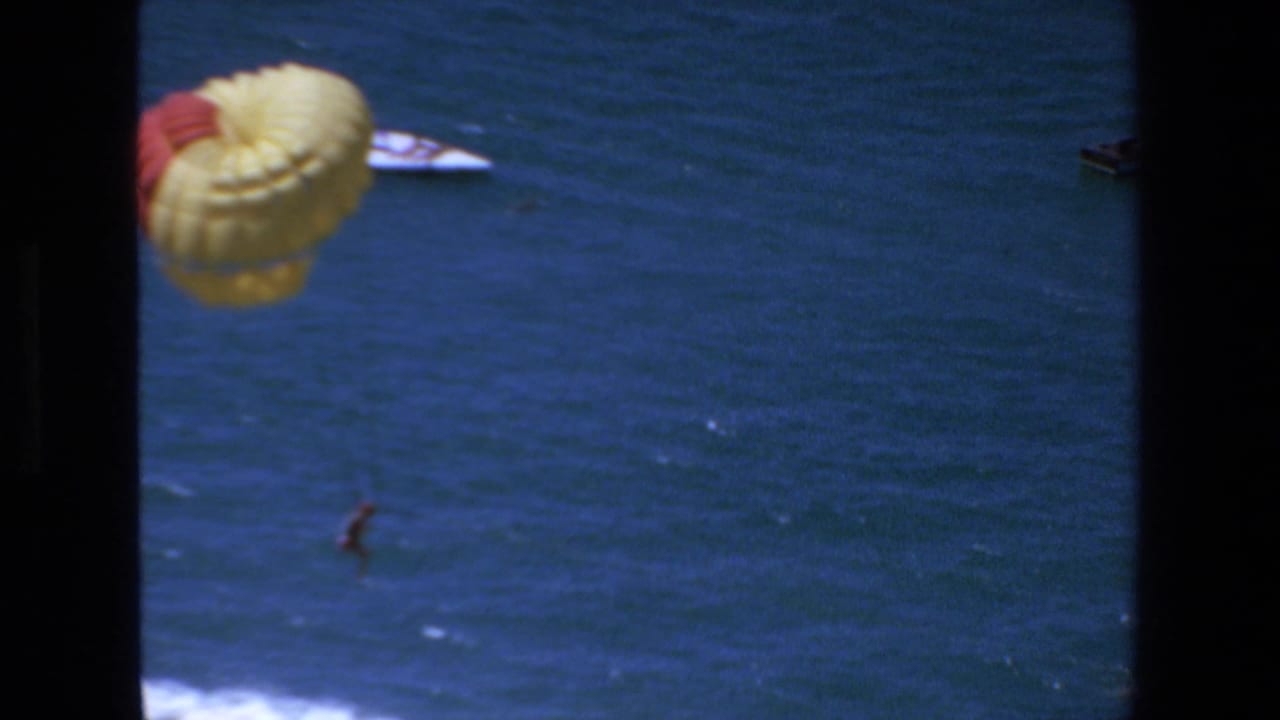 1972 Mazatlan home-movie frame showing a yellow-and-red parasail over deep blue ocean water, with a distant parasailer above white surf and dark 8mm frame edges.