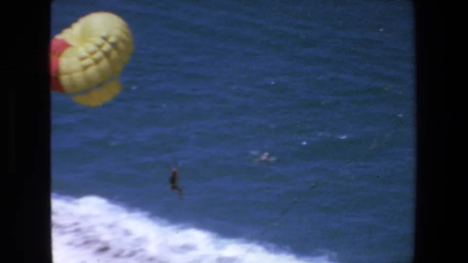 1972 Mazatlan 8mm frame showing a yellow-and-red parasail at left, the parasailer small against blue water, and white surf rolling across the lower edge.