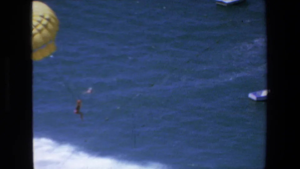 1972 Mazatlan home-movie frame with the parasailer small over open blue water, white surf below, and a small motorboat partly visible near the right edge.