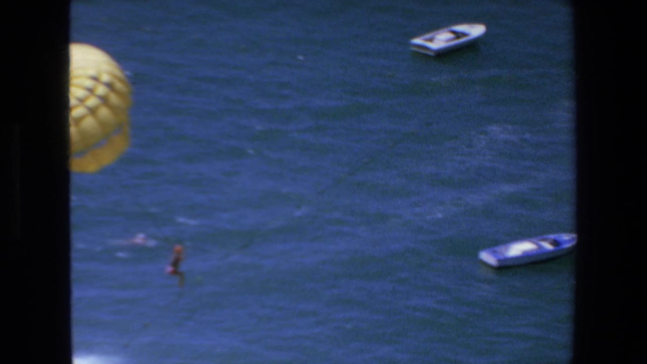 1972 Mazatlan home-movie frame showing a distant parasailer suspended below a yellow parachute over blue ocean water, with small boats visible around the activity.