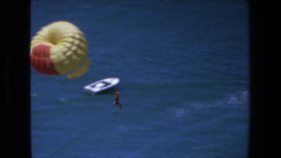 1972 Mazatlan home-movie frame showing the parasail canopy nearer the left side, a rider hanging below, blue ocean water, and a small white boat beneath.