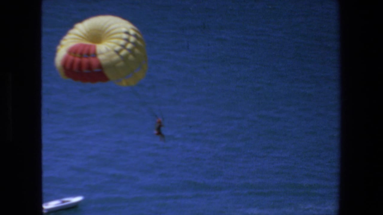 1972 Mazatlan 8mm frame showing a red-and-yellow parasail canopy over deep blue water, with a distant person hanging below and a small white boat partly visible near the lower left.