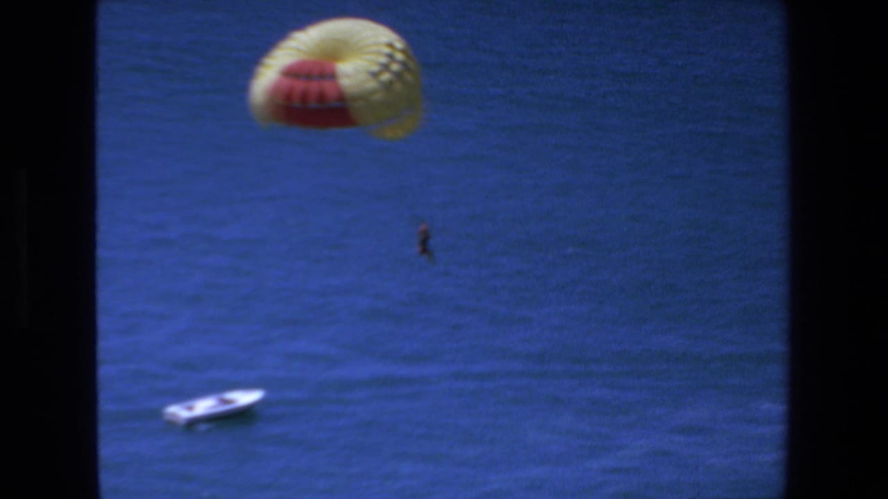 Parasail canopy high over blue water with a distant suspended person and small white boat below.