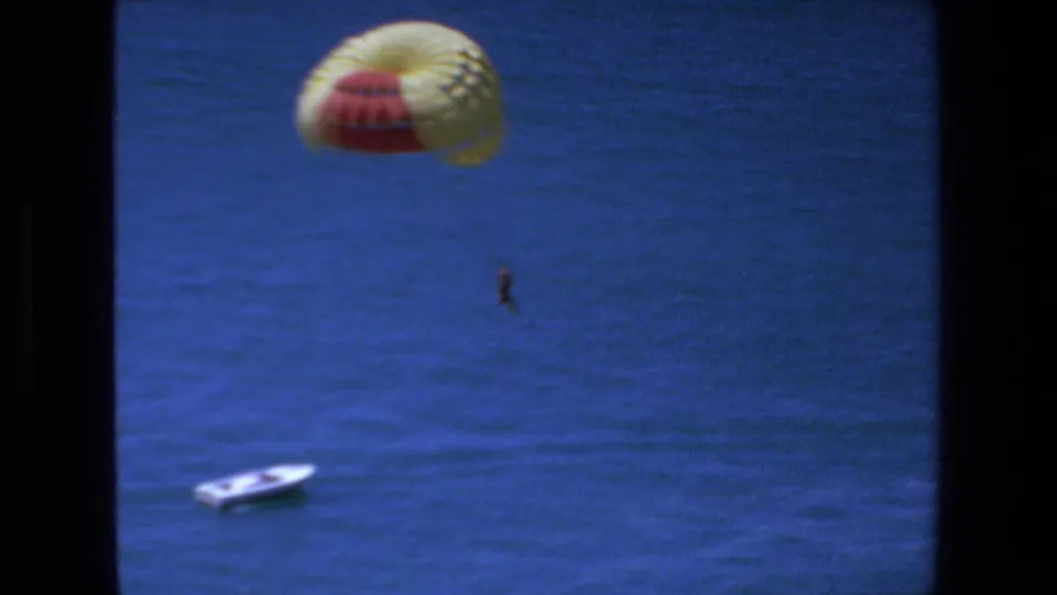 1972 Mazatlan 8mm frame with the red-and-yellow parasail canopy near the top of the shot, a small suspended figure below it, and a white boat on the ocean beneath.